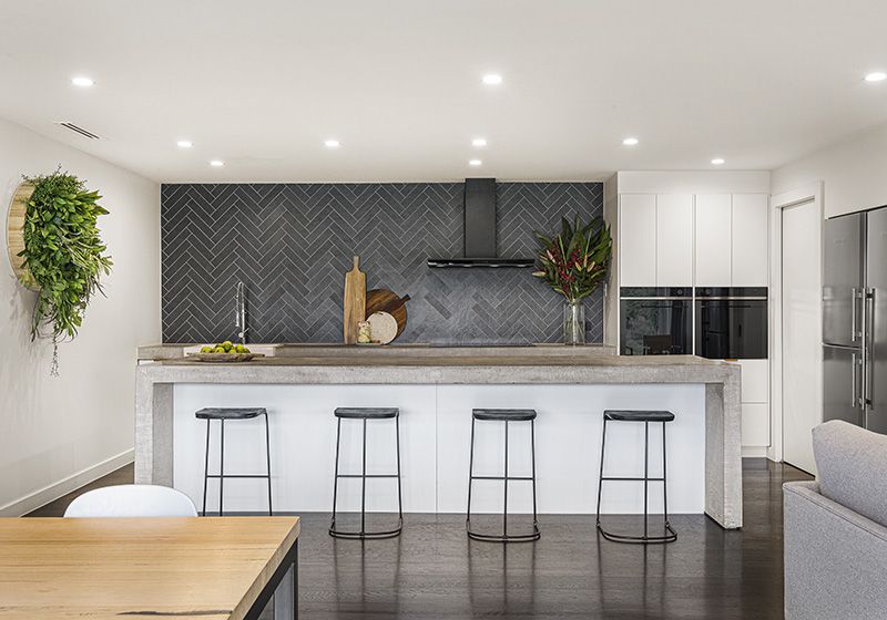 Cocina abierta con isla y cuatro taburetes. En el frente una campana decorativa y una pared con azulejo negro.